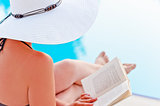 Girl in a hat reading a book near the pool, sitting in a lounge chair