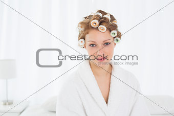 Beautiful woman in bathrobe and hair curlers sitting on bed