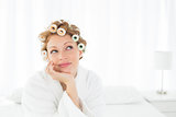 Thoughtful woman in bathrobe and hair curlers sitting on bed