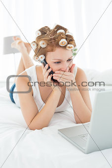 Shocked woman in hair curlers using cellphone and laptop in bed