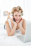 Woman in hair curlers using cellphone and laptop in bed