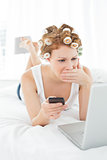 Shocked woman in hair curlers with cellphone looks at laptop in bed