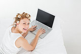 Cheerful woman in hair curlers using laptop in bed