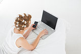 Woman in hair curlers with cellphone using laptop in bed