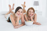Two happy female friends with popcorn bowl lying in bed