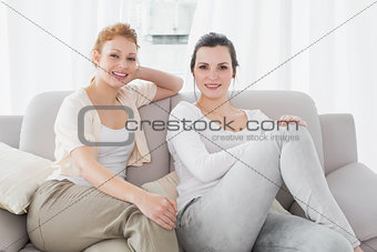 Two smiling female friends sitting on sofa in living room