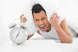 Man covering ears with pillow and shouting with alarm clock in foreground