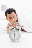 Sleepy man in bed extending hand to alarm clock