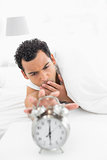 Sleepy man in bed extending hand to alarm clock