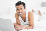 Smiling casual young man using laptop in bed