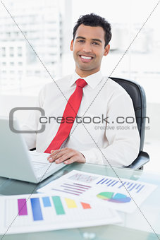 Smiling businessman with laptop and graphs sitting at office
