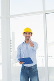 Architect in hard hat with clipboard gesturing thumbs up in office