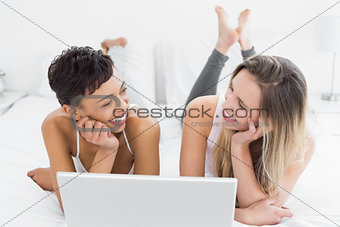 Cheerful female friends using laptop in bed