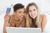 Happy relaxed female friends doing online shopping in bed