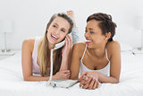 Cheerful young female friends using phone in bed