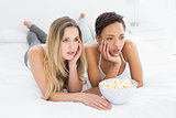 Serious female friends with popcorn bowl lying in bed