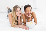 Cheerful female friends with popcorn bowl lying in bed