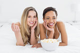 Cheerful female friends with popcorn bowl lying in bed