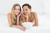 Happy female friends with popcorn bowl lying in bed
