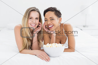 Happy female friends with popcorn bowl lying in bed