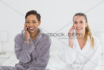 Female friends in bathrobes using phones on bed