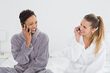 female friends in bathrobes using phones on bed