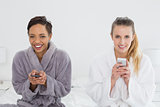 Female friends in bathrobes text messaging on bed