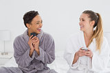 Cheerful friends in bathrobes text messaging on bed