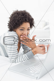Shocked woman with coffee cup using laptop in bed