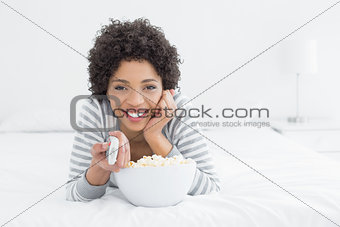 Smiling woman with remote control and popcorn bowl in bed