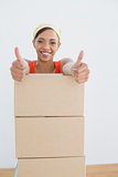 Woman gesturing thumbs up with stack of boxes in a new house