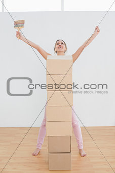 Woman with boxes and paintbrush raising hands in new house