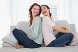 Cheerful young female friends sitting back to back in the living room