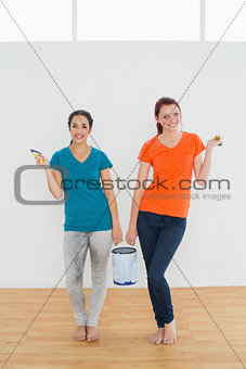 Smiling female friends with brushes and paint can in new house