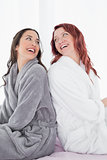 Happy female friends in bathrobes sitting back to back on bed