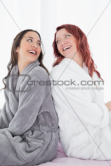 Happy female friends in bathrobes sitting back to back on bed