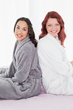 Female friends in bathrobes sitting back to back on bed
