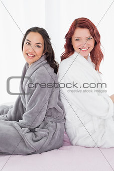 Female friends in bathrobes sitting back to back on bed