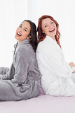 Cheerful friends in bathrobes sitting back to back on bed