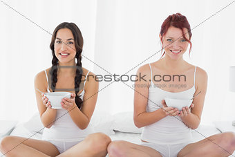 Smiling young female friends with bowls sitting on bed