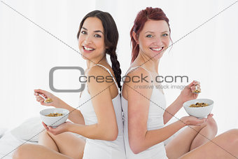 Side view of smiling friends with bowls sitting on bed