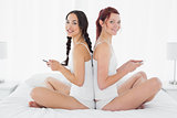 Female friends sitting back to back with cellphone on bed