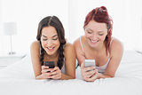 Smiling female friends text messaging on bed