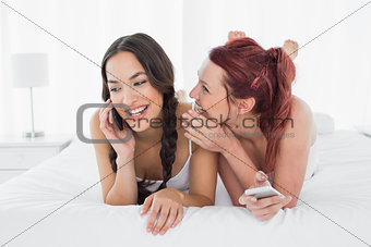 Woman whispering into cheerful friends ear while on call in  bed