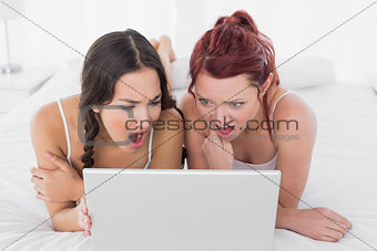 Shocked female friends looking at laptop in bed