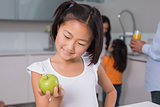 Smiling young girl holding apple with family in at kitchen