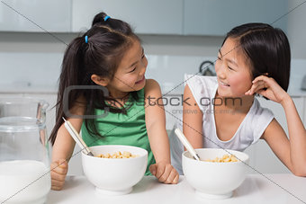 Two smiling young girls sitting with cereal bowls in kitchen