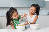 Two happy young girls pouring milk in bowl in kitchen