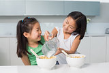 Two happy young girls pouring milk in bowl in kitchen