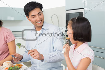 Father watching little girl eat food with a fork in kitchen